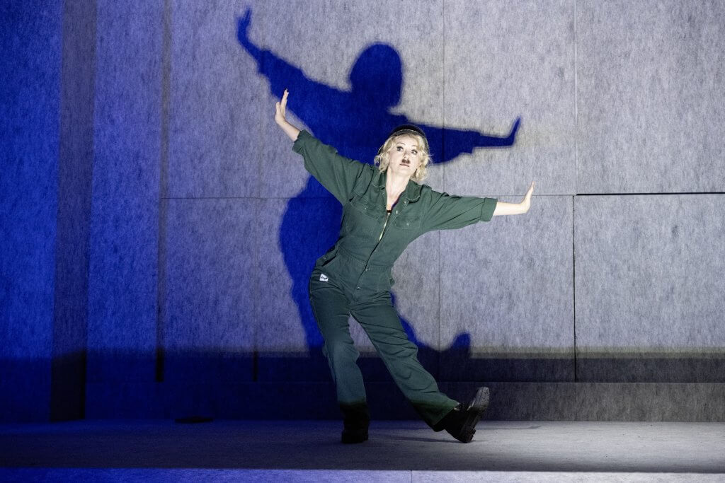 Eine Frau in einem grünen Overall steht in einer Show-Pose auf der Bühne.