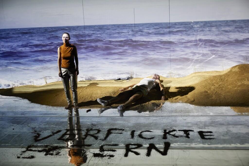 Eine Person steht, eine andere liegt auf einer Bühne, die an einen Strand erinnert.