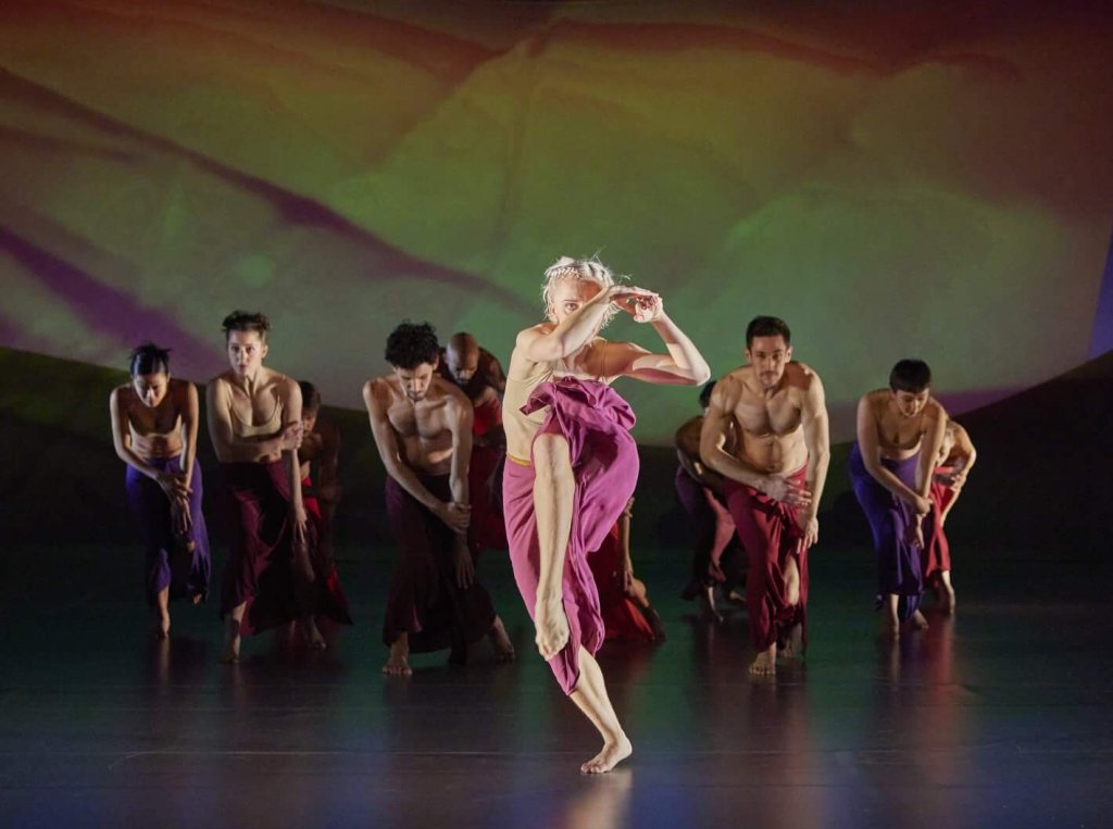 Eine Person in einem violetten Kleid tanzt ekstatisch vor einer Gruppe tanzender Personen, die sich gebückt in zwei Reihen bewegen.