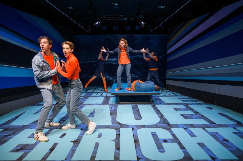 Zwei Personen in orangenen Oberteilen und Jeans stehen in Tanzhaltung auf einer blauen Bühne. Im Hintergrund gestikulieren weitere Menschen.