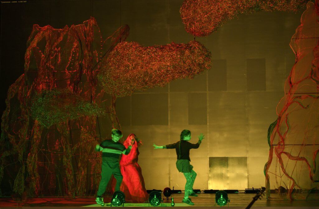 Auf einer Bühne in grünen und roten Licht tanzen eine Frau und zwei Kinder.