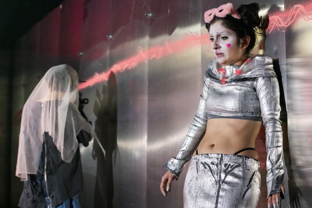 Eine Frau in silberglänzernder Kleidung drückt sich an eine Metallwand. Eine Person unter einem Schleier filmt sie.