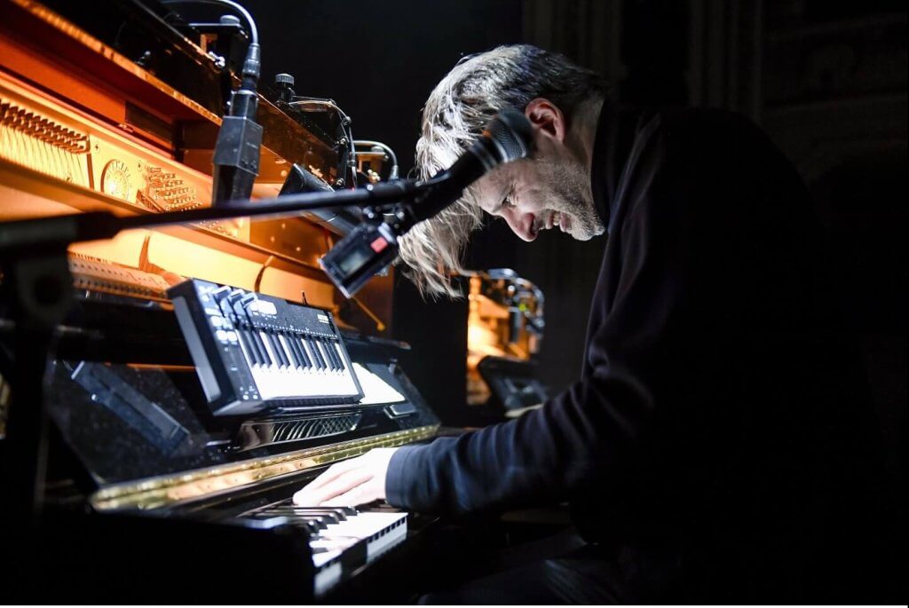 Ein Mann sitzt an einem Klavier, an dem zusätzlich Mikrofone und ein Keyboard angebracht sind.