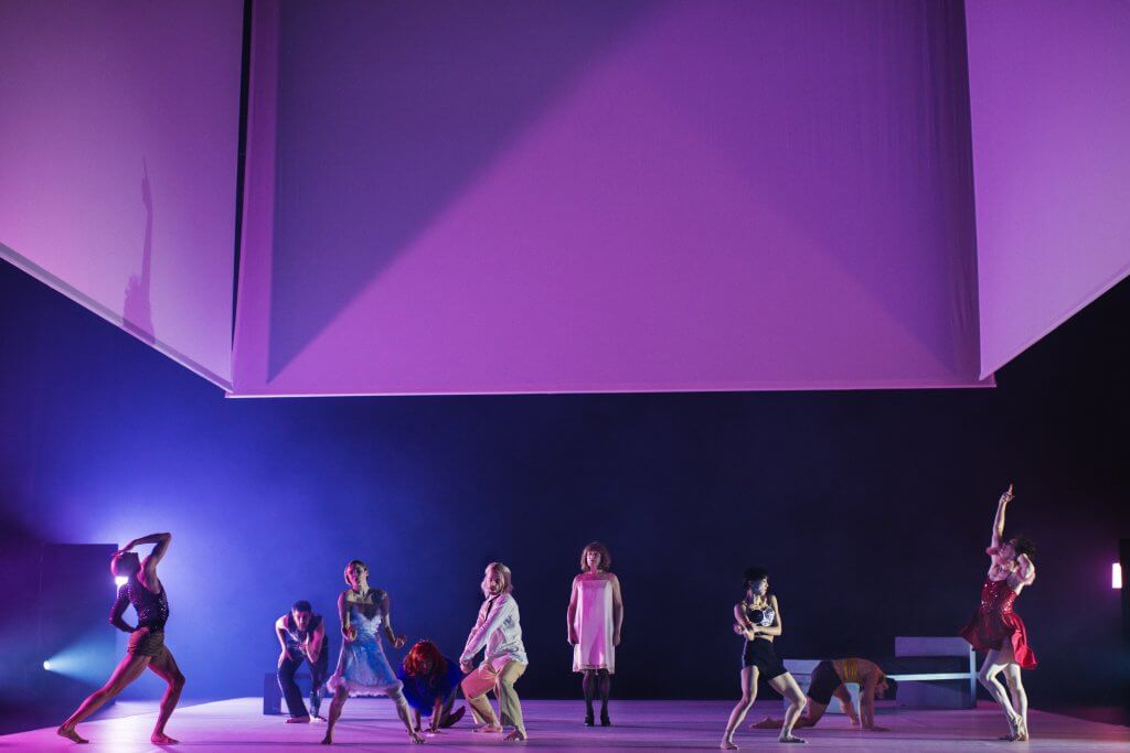 Wände schweben über der Bühne und werden rosa und blau angeleuchtet. darunter stehen und tanzen mehrere PErsonen in einer Reihe.