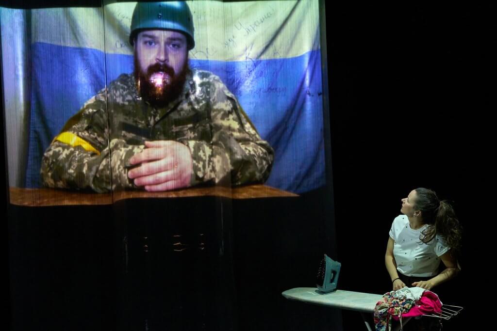 Im Hintergrund die Projektion einer Person in Soldatenuniform mit Helm vor ukrainischer Flagge. Schräg rechts davor eine Frau am Bügelbrett, die zur Projektion aufschaut