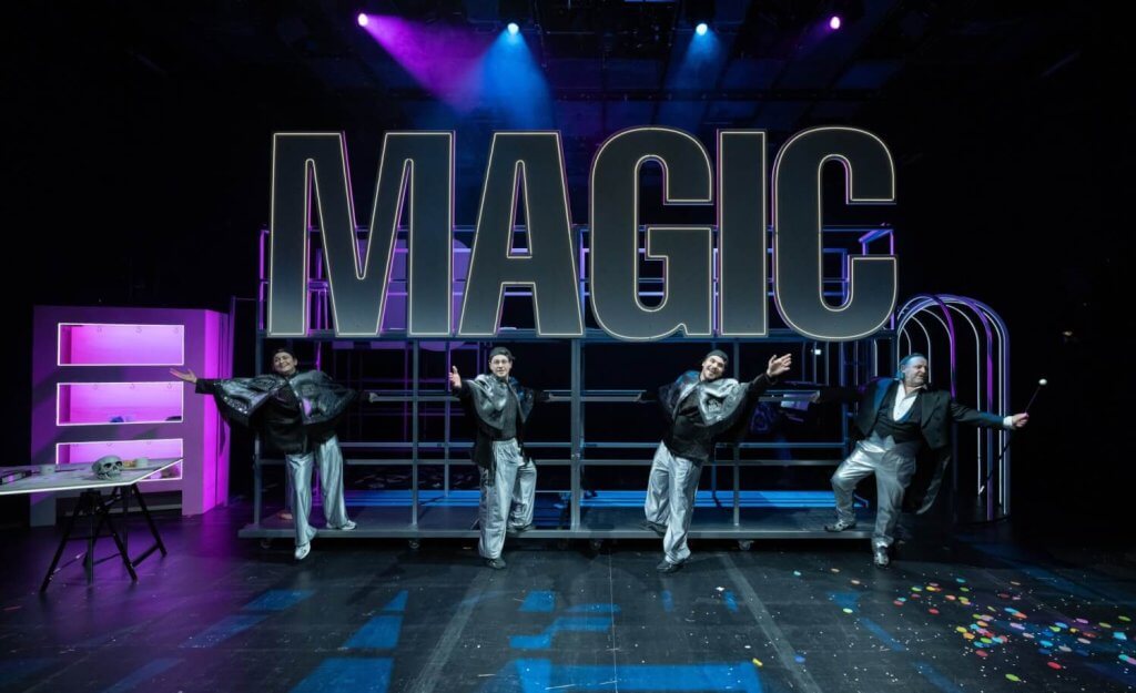 Vier Personen in schwarz-silbernen Show-Anzügen posieren mit nach vorn gestreckten Armen vor den riesigen Lettern "Magic".