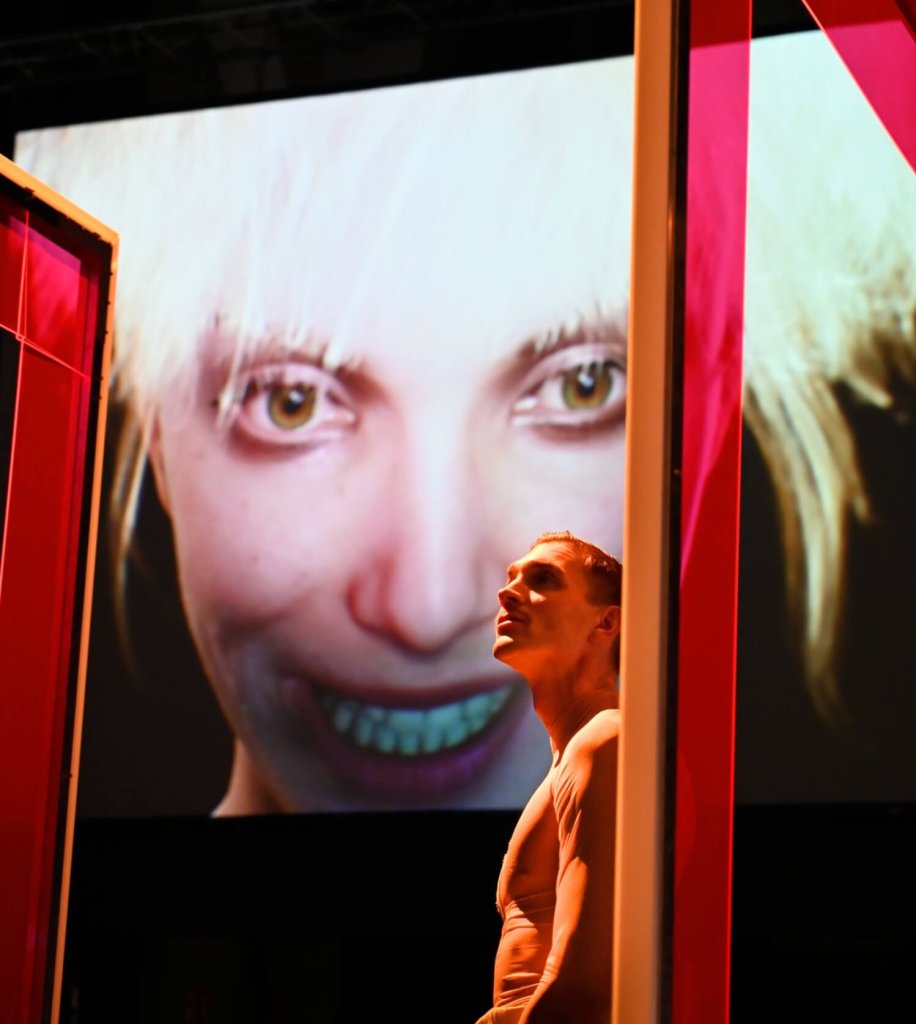 zwischen zwei menschengroßen roten Rahmen im Hintergrund ein großer Bildschirm mit einem Avatargesicht mit kurzen blonden Haaren, davor Ensemlbesänger Simon Mantei als Julian Zapp, schaut nach links oben