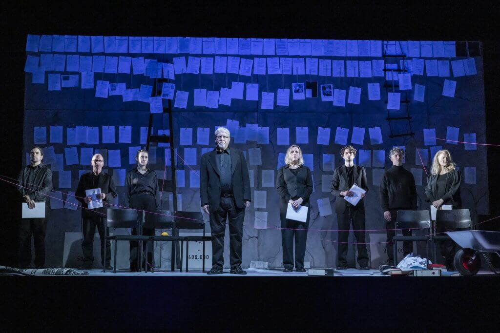 Menschen in schwarzer Kleidung stehen auf einer blau leuchtenden Bühne in einer Reihe vor einer Wand voller Zettel.