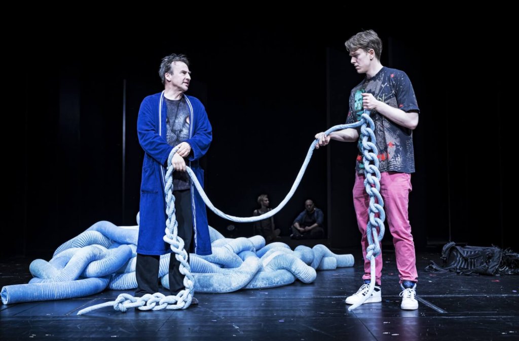 Zwei Männer stehen auf einer Bühne und halten jeweils das Ende eines Seils, das schlaff zwischen ihnen hängt.