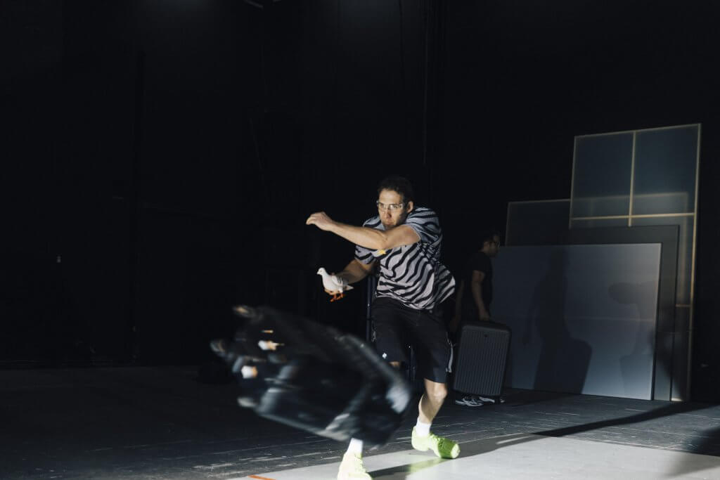 Schauspieler in Zürich in der Inszenierung von Williams' „Moise und die Welt der Vernunft“ mit Brille und Taubenfigur in der Hand wirf mit konzentriertem Blick einen Koffer von sich schräg nach vorne links
