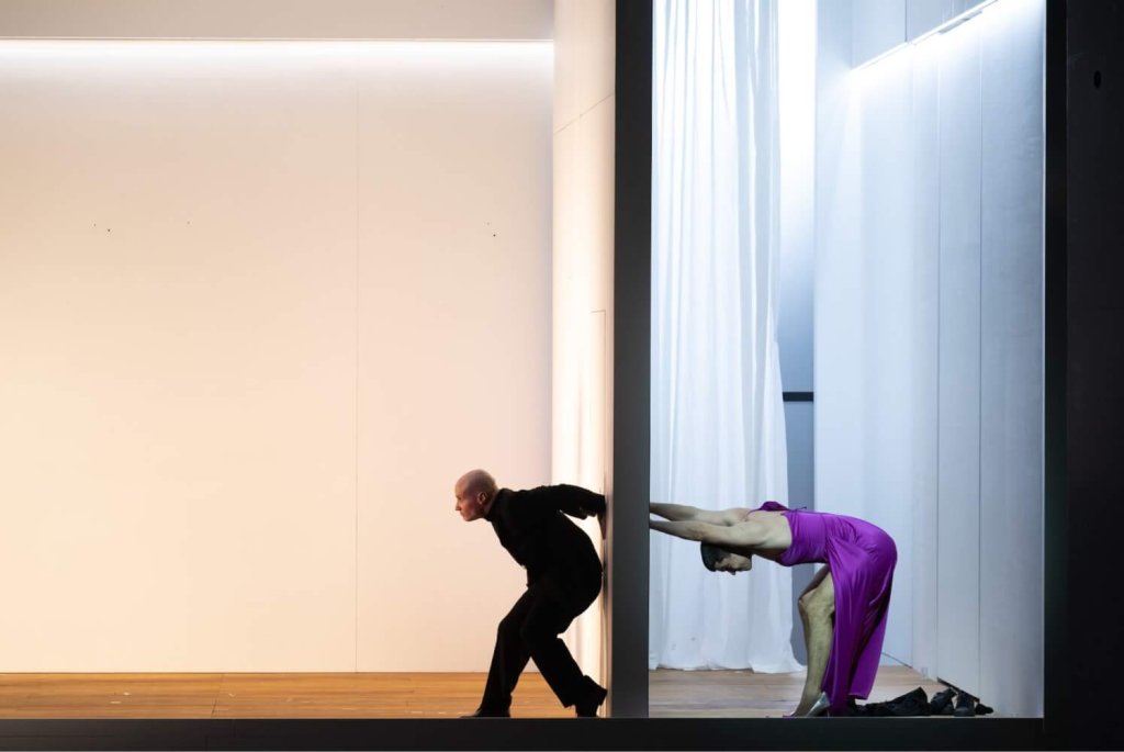 Eine Frau in einem violetten Satinkleid drückt mit beiden Händen gegen eine Wand, auf der anderen Seite drückt ein Mann im Anzug mit dem Rücken dagegen.