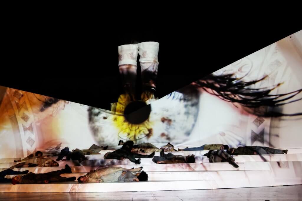 Eine Projektion eines riesigen Auges, davor liegen mehrere Personen flach auf der Bühne.