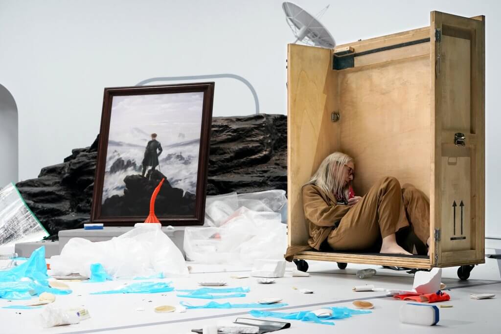 Eine Person in einem beigen Overall kauert in einer Art Holzkiste, daneben steht das Bild "Wanderer über dem Nebelmeer", ringsherum liegt blaues und weißes Plastik.