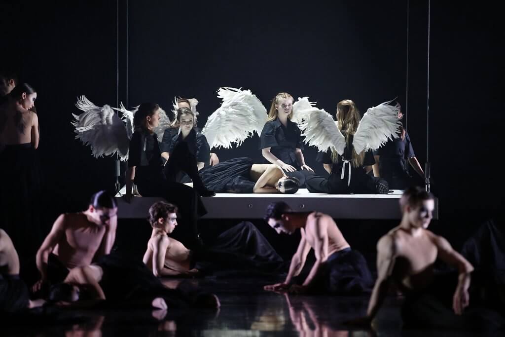 Frauen mit weißen Federflügeln hocken auf einer Bühne, davor bewegen sich weitere Personen in Schwarz auf dem Boden.