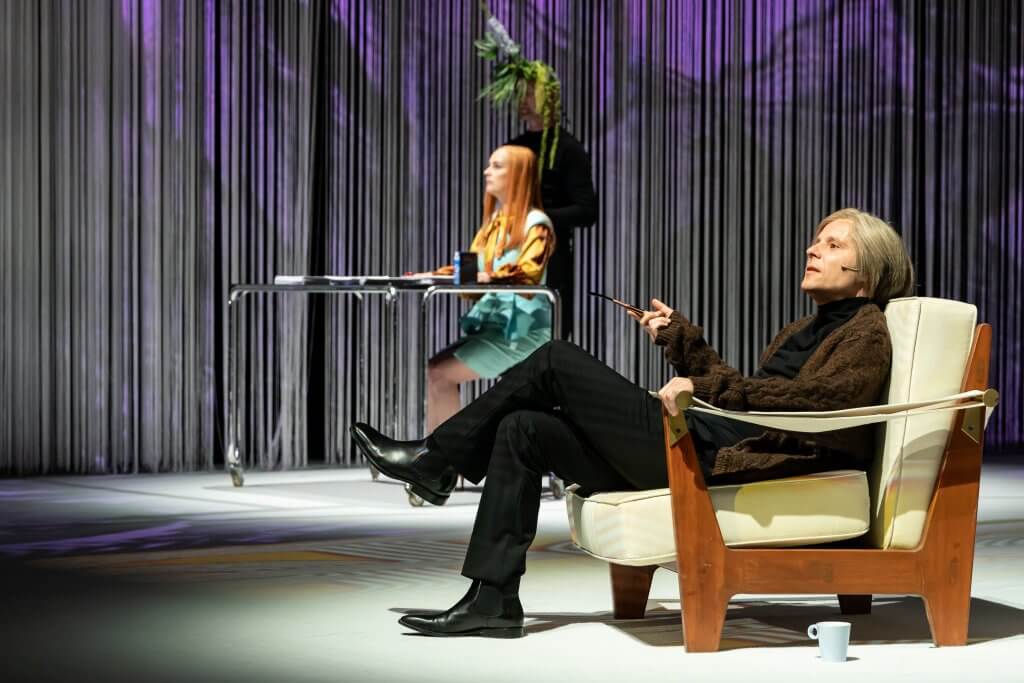 Eine Person sitzt auf einem Sessel mit Holz-Rahmen, dahinter eine weitere Person an einem kleinen Glastisch, im Hintergrund ein Fadenvorhang.