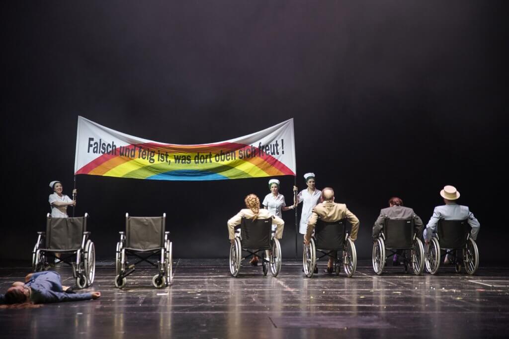 Menschen in Rollstühlen in einer Reihe vor einem Banner mit Regenbogen und Schriftzug "Falsch und feit ist, was dort oben sich freut!