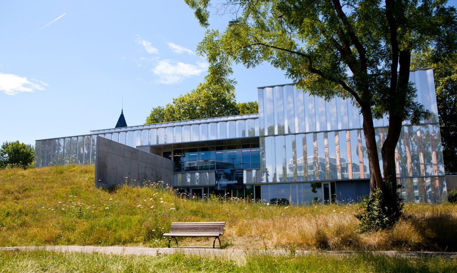 Theater Reutlingen Die Tonne Man sieht das Theater Reutlingen Die Tonne. Im Vordergrund rechts steht ein Baum, in der Mitte vor dem Gebäude steht inmitten von Gras eine Sitzbank.