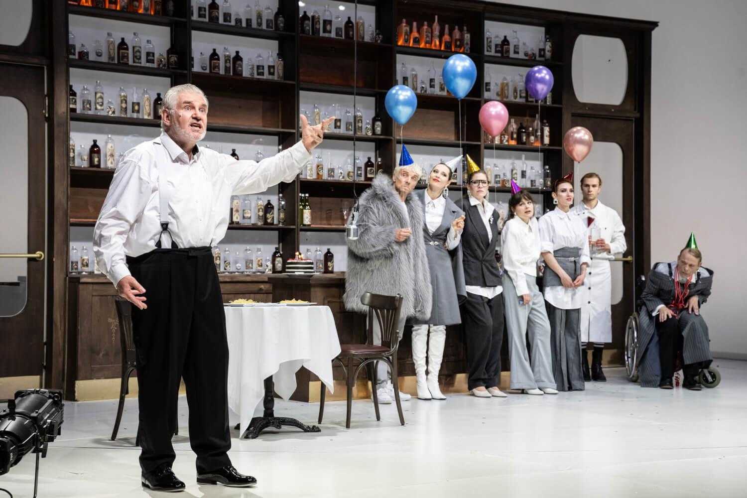 Vorne links steht ein älterer Herr in Hemd Abendkleidung. Im Hintergrund steht eine Partygesellschaft mit Partyhüten und Luftballons.