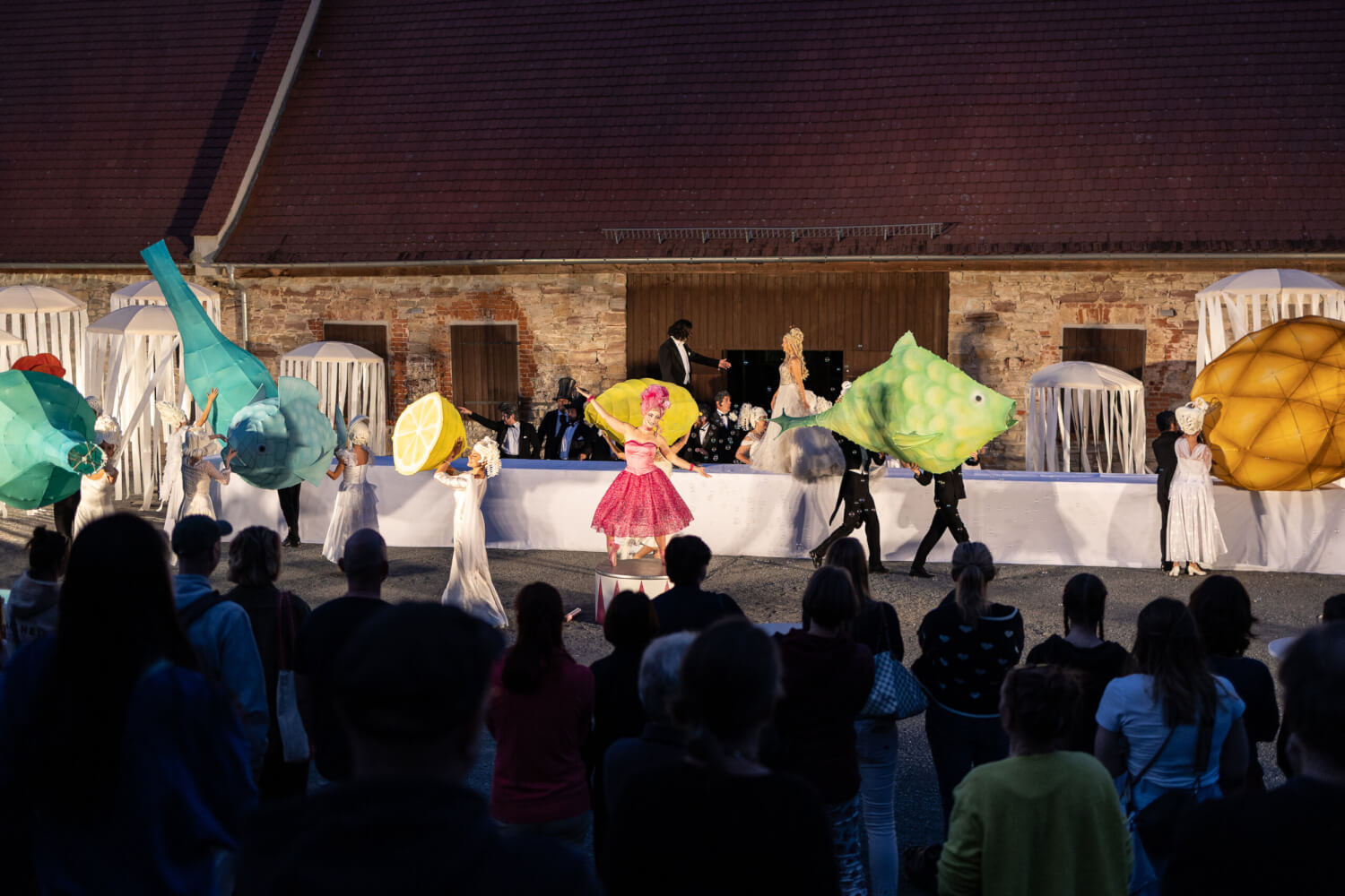 Im Vordergrund befindet sich das im Schatten stehende Publikum. Auf der Bühne findet Orpheus und Eurydikes Hochzeit statt. Ein langer gedeckter Tisch mit zahlreichen bunt gekleideten Gästen ist zu sehen. Sie tragen übergroße Fische, Flaschen und Früchte zu Tisch.