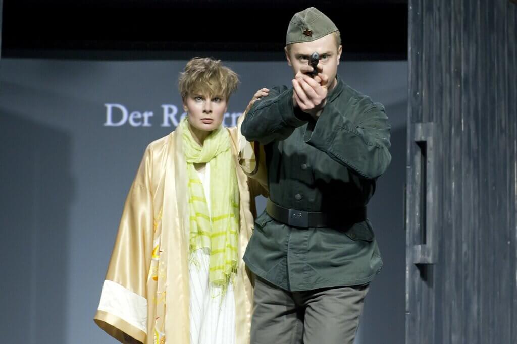 rechts ein Darsteller in Soldatenuniform mit Waffe, die in die Kamera zielt, links daneben eine Frau im Morgenmantel und mit grünem Halstuch