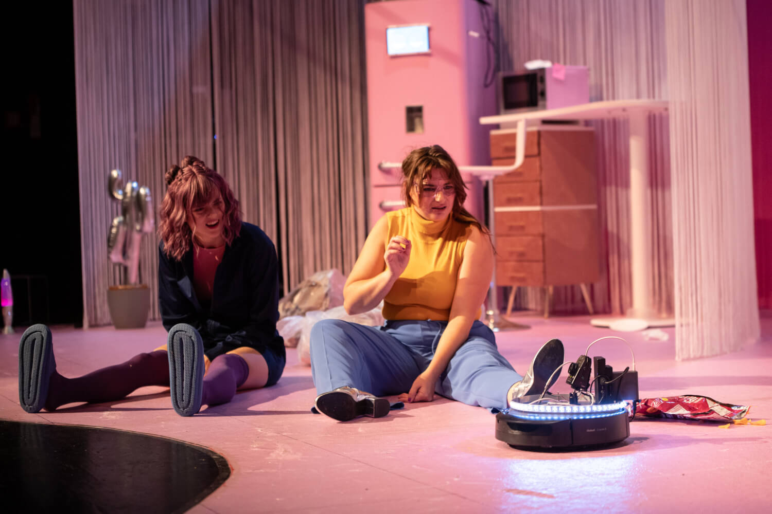 Cuba (Billie Barleben) und Fanny (Marie-Luis Kießling) sitzen in ihrer WG-Küche auf dem Boden mit Volker dem Staubsauger-Roboter.
