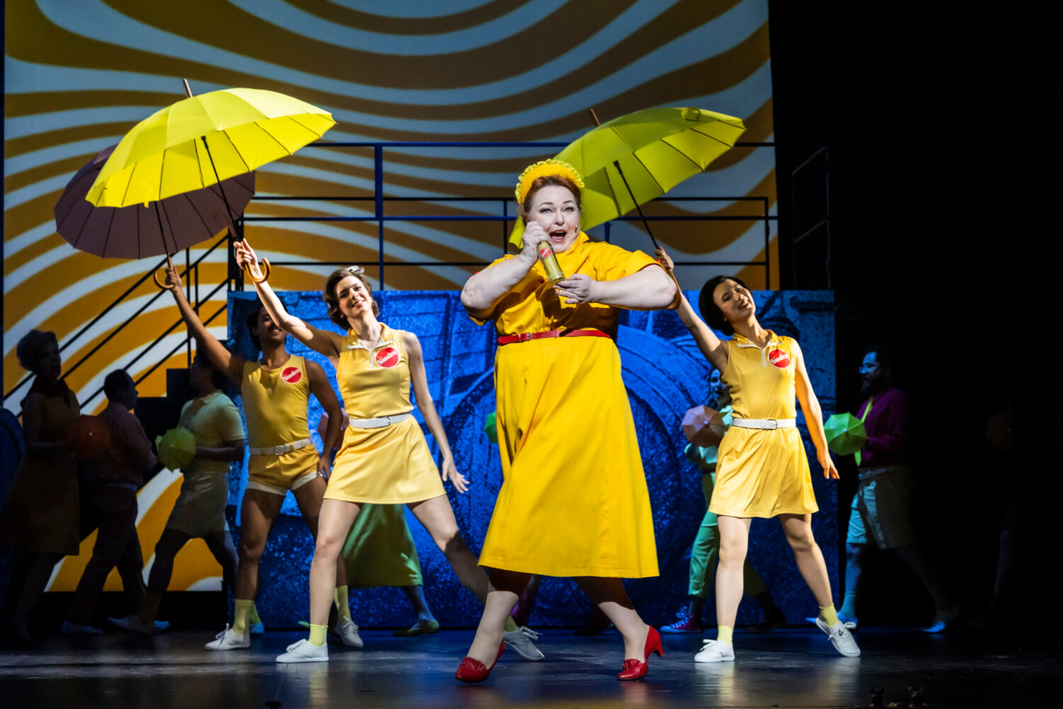 Aufführungsfoto von „Das Glück ist eine Orange. Sinalco - das Musical!“ am Landestheater Detmold. Eine Frau im gelben Kleid hält eine Sinalcoflasche in den Händen und geht singend im Kreuzschritt über die Bühne. Im Hintergrund recken drei Tänzer:innen in gelber Kleidung und einem roten Sinalco-Logo auf der Brust, gelbe Schirme in die Höhe.