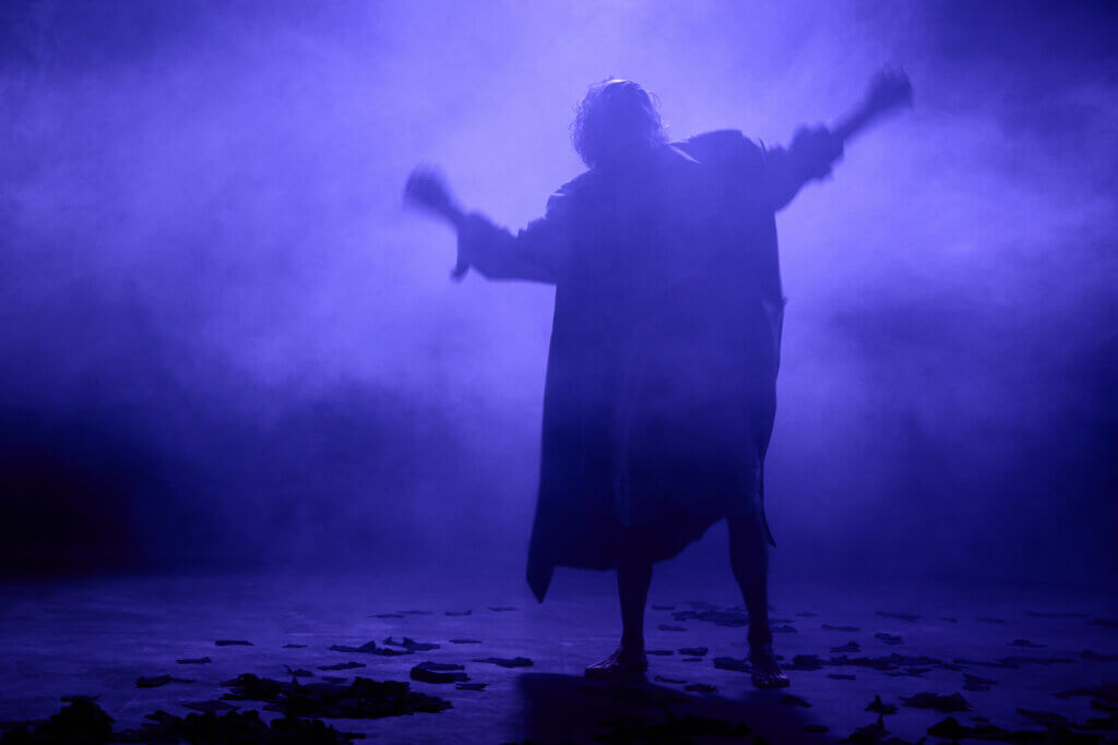 Szenenfoto aus „Die Erbschaft“ von Jasmin Meindl und Christian Muggenthaler am Landestheater Dinkelsbühl. Eine Frau reckt die Arme nach oben, den Kopf in den Nacken geworfen. Ihre Silhoutte ist in blau-lila Licht und Nebel getaucht.