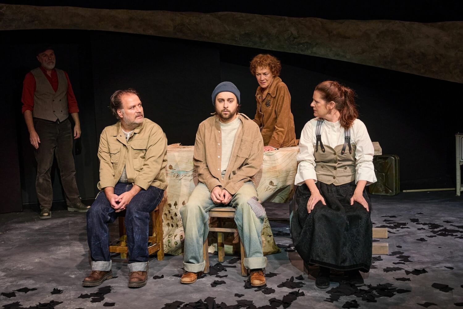 Szenenfoto aus „Die Erbschaft“ von Jasmin Meindl und Christian Muggenthaler am Landestheater Dinkelsbühl. Zwei Männer und eine Frau sitzen auf drei Holzschemeln. Dahinter lehnt eine Frau an einer Bank. Links im Hintergrund steht ein Mann.