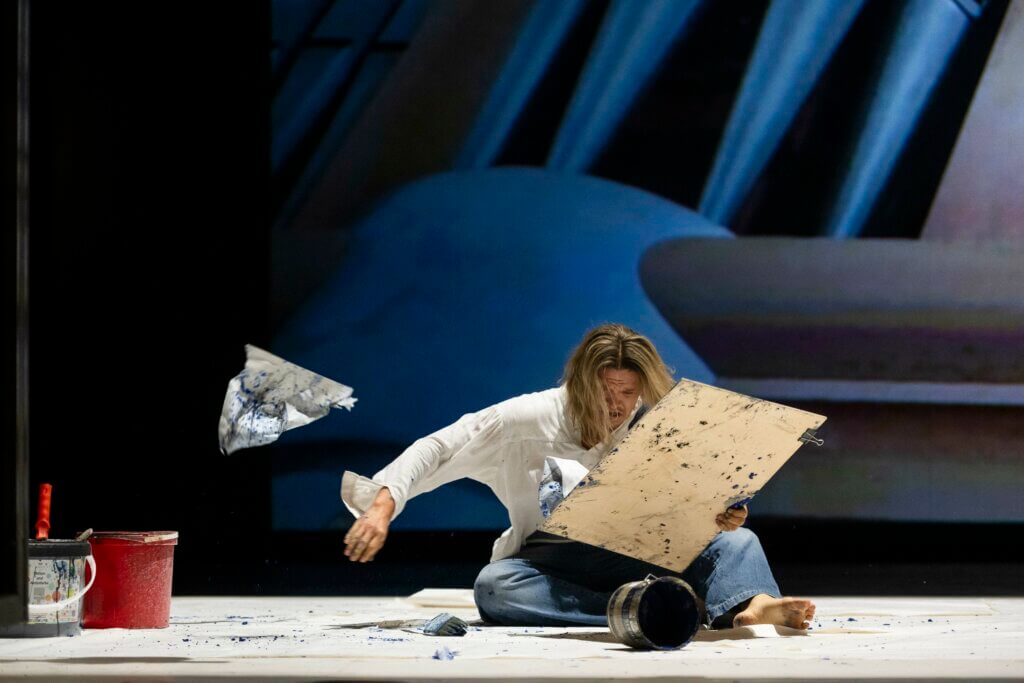 Aufführungsfoto von „Die Nibelungen – Rang und Drang“ von Marcel Luxinger und Ivana Sokola. Ein Mann sitzt auf dem Boden, eine Leinwand in der linken Hand, wirft er ein von der Leinwand zerrissenes Stück Papier von sich. Vor ihm ein umgefallender Farbeimer.