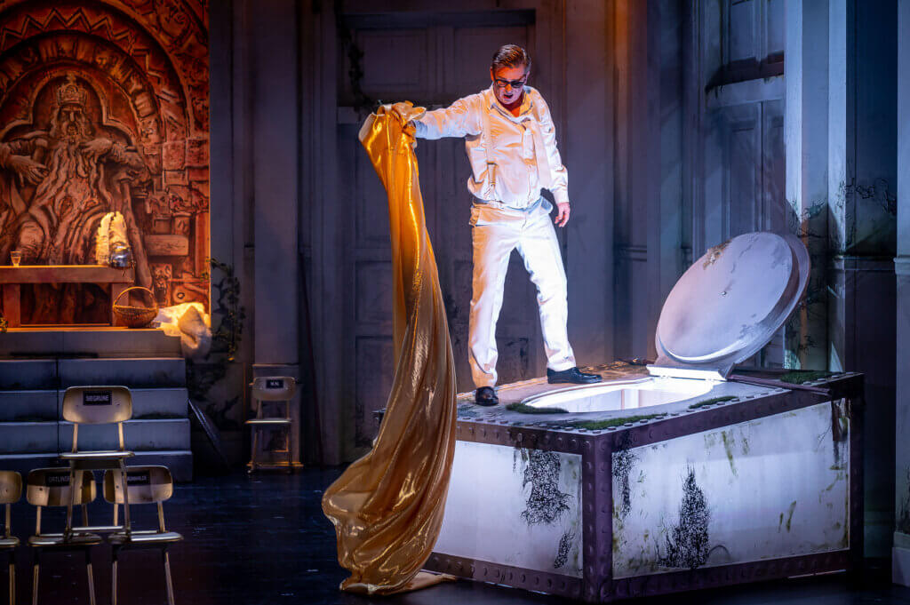 Aufführungsfoto von „Die Walküre“ von Richard Wagner am Harztheater in Halberstadt. Ein Sänger steht auf einem weiß erleuchtetem Quadrat, dessen Luke geöffnet ist. Er hält einen langen, goldfarbenen Stoff in der rechten Hand.