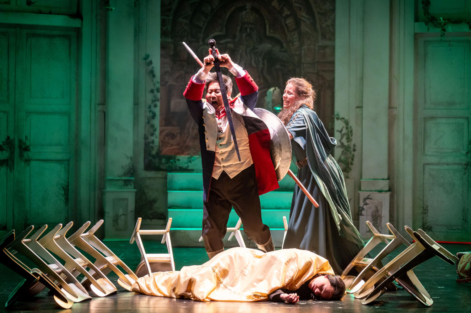 Aufführungsfoto von „Die Walküre“ von Richard Wagner am Harztheater in Halberstadt. Ein Sänger holt aus, um mit einem Schwert auf die am Boden liegende Frau einzustechen. Eine Fraz hinter ihm versucht ihn verzweifelt davon abzuhalten.