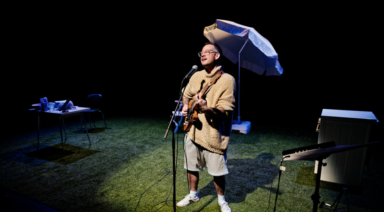 Henri Maximilian Jakobs steht im Spotlight mit umgehängter Gitarre singend vor einem Mikrofon, dahinter sieht man einen Sonnenschirm
