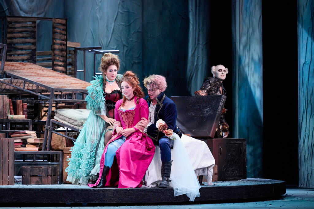 Aufführungsfoto von „Der tollste Tag“ von Johanna Donderer am Gärtnerplatztheater in München. Zu sehen sind zwei Sängerinnen und ein Sänger im Vordergrund sitzend. Im Hintergrund ein Sänger mit weiß geschminktem Gesicht und schwarz umrandeten Augen.