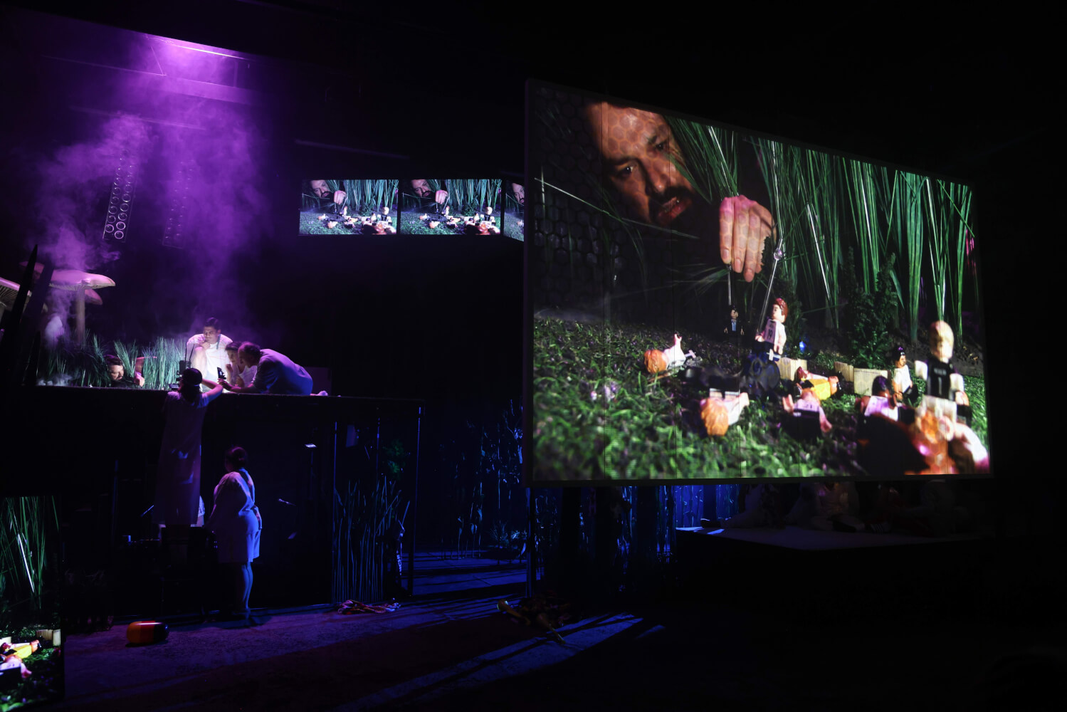 Szenenfoto aus „Der Zauberer von Öz – Eine Fußballtragödie“ von Akın Emanuel Şipal am Theater Bremen. Die Bühne ist in dunkles, blau-lila Licht getaucht. Vorne rechts ein großer Bildschirm, auf dem der Kopf eines Mannes zu sehen ist hinter Grasbüscheln und vorne Lego-Figuren.