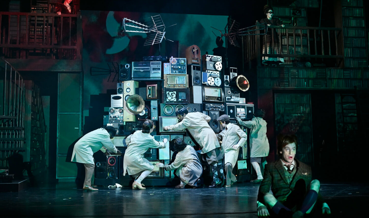 Das Ensemble umarmt einen Berg aus aufgetürmten Lautsprechern. Jan-Peter Kampwirth sitzt als Hildegunst von Mythenmetz vorne auf dem Bühnenboden und schaut mit Schrecken ins Publikum.