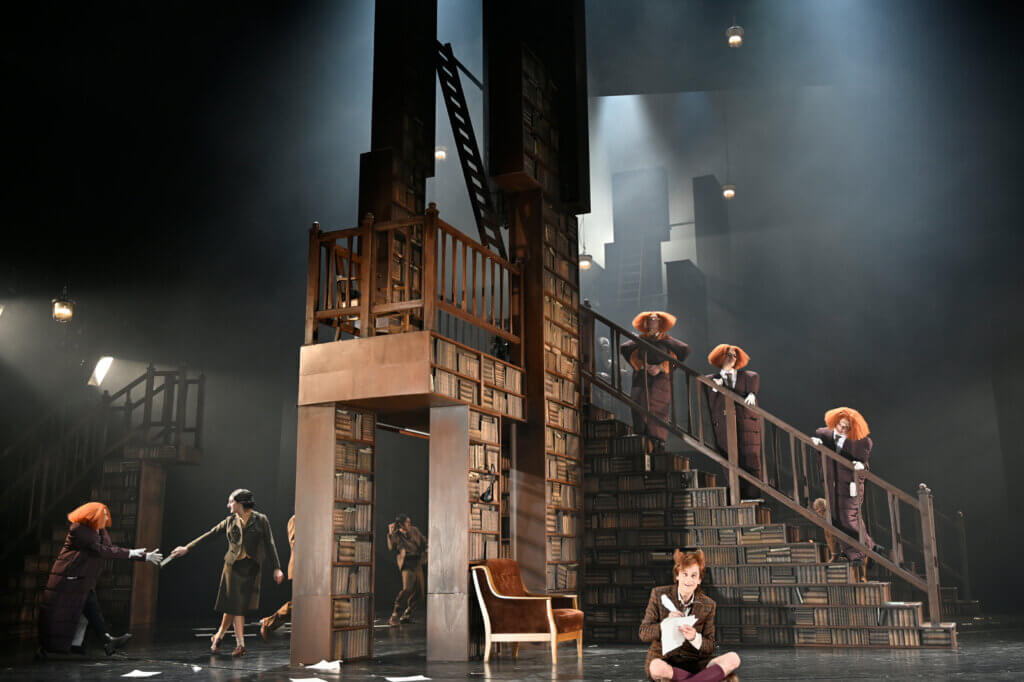 Auf der Bühne steht eine große mehrstöckige Bibliothek. Im Vordergrund sitzt Jan-Peter Kampwirth im Schneidersitz auf dem Boden.