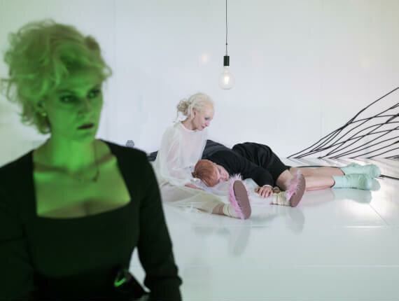 EIne Frau mit hochtoupierten Haaren, im Hintergrund hält eine Frau in Weiß eine Person im Schoß.