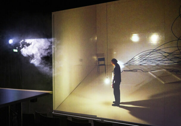 Eine Person steht alleine auf einer Bühne mit einzelnen Lichtern und mehreren unordentlichen Kabeln an der Wand.