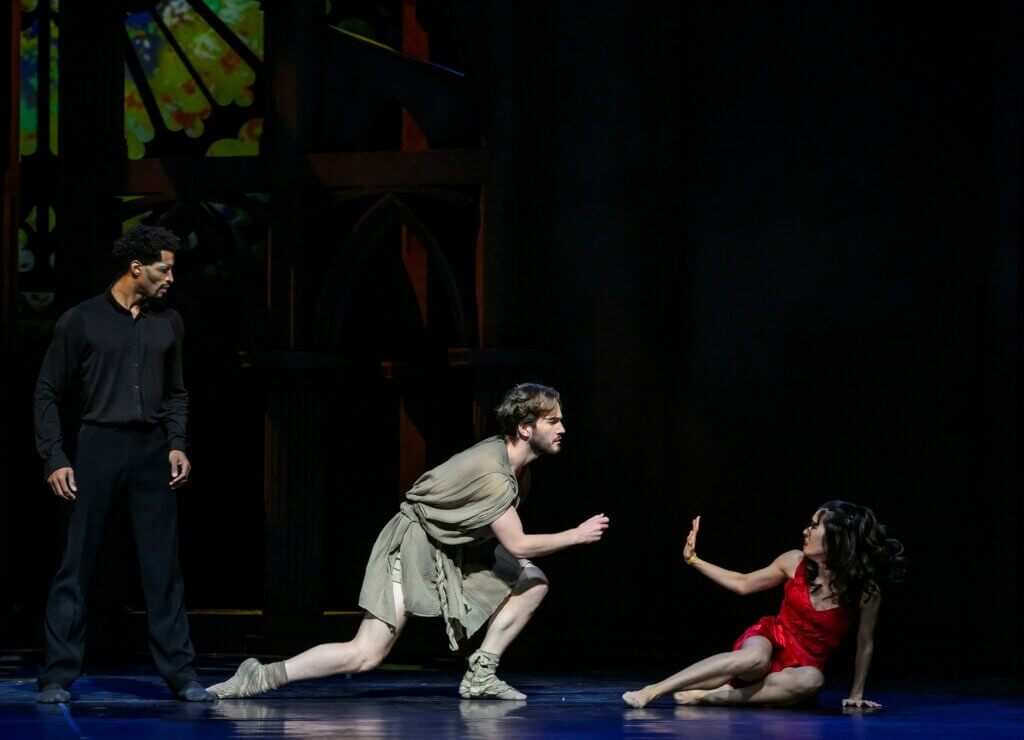 Esmeralda sitzt rechts auf dem Bühnenboden und streckt abwehrend ihre Hand in Richtung Quasimodo. Der versucht in Kniebeugenhaltung mit ihr in Kontakt zu treten. Hinter ihm steht Frollo, der alles beobachtet.