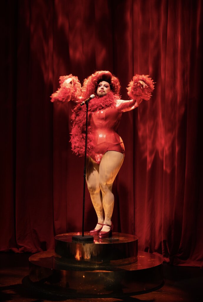 eine Person im roten Drag-Outfit vor einem Mikrofon seitlich von links mit erhobenen Armen