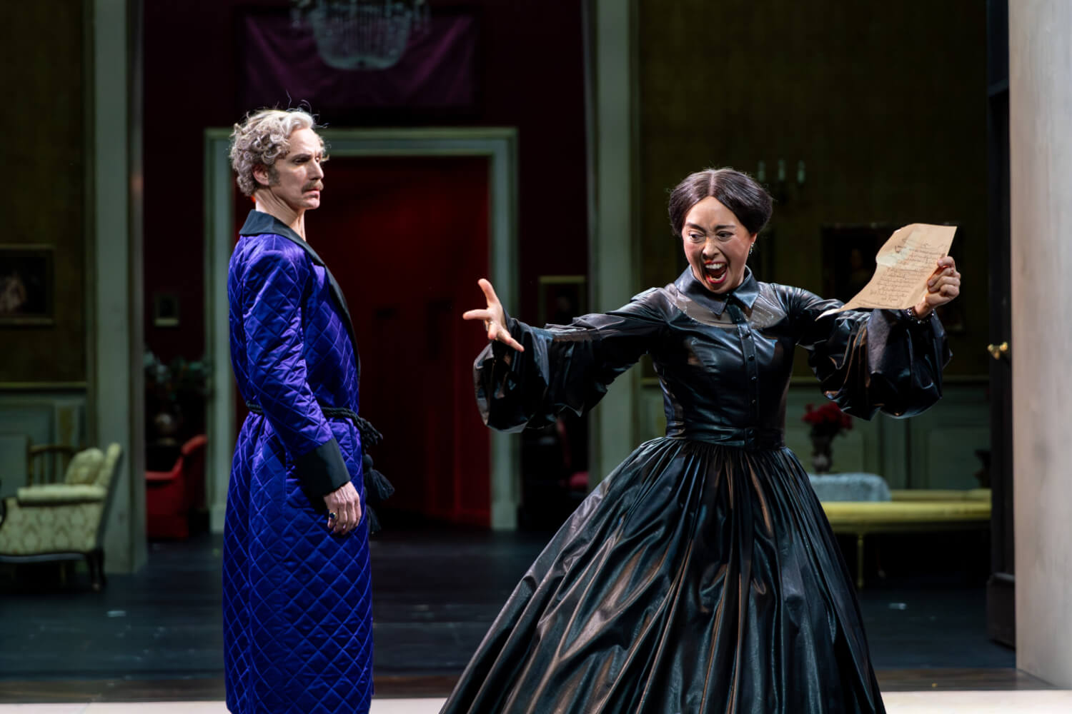 Aufführungsfoto von „Il Gattopardo“ von Giuseppe Tomasi di Lampedusa am Schauspielhaus Zürich. Eine Frau im schwarzen Kleid hält in der linken Hand einen Brief, die rechte Hand spreizt sie von sich und schreit. links neben ihr steht ein Mann im blau-samtenen Bademantel und guckt sie skeptisch an.