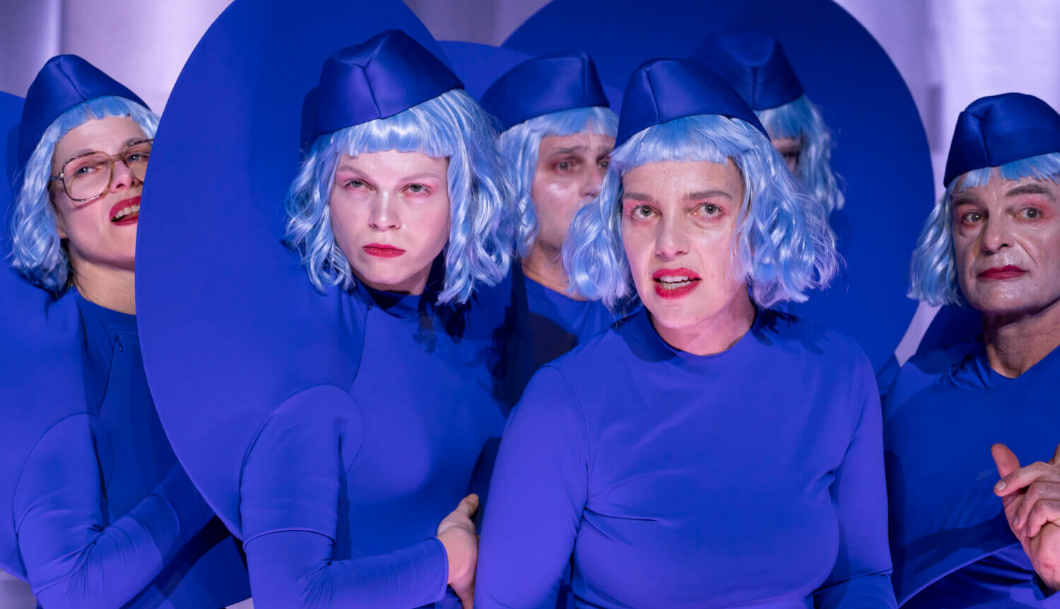 Das Ensemble steht Ganzkörperanzügen und kurzen, gelockten Perrücken in einer Gruppe und schaut misstrauisch nach vorn.