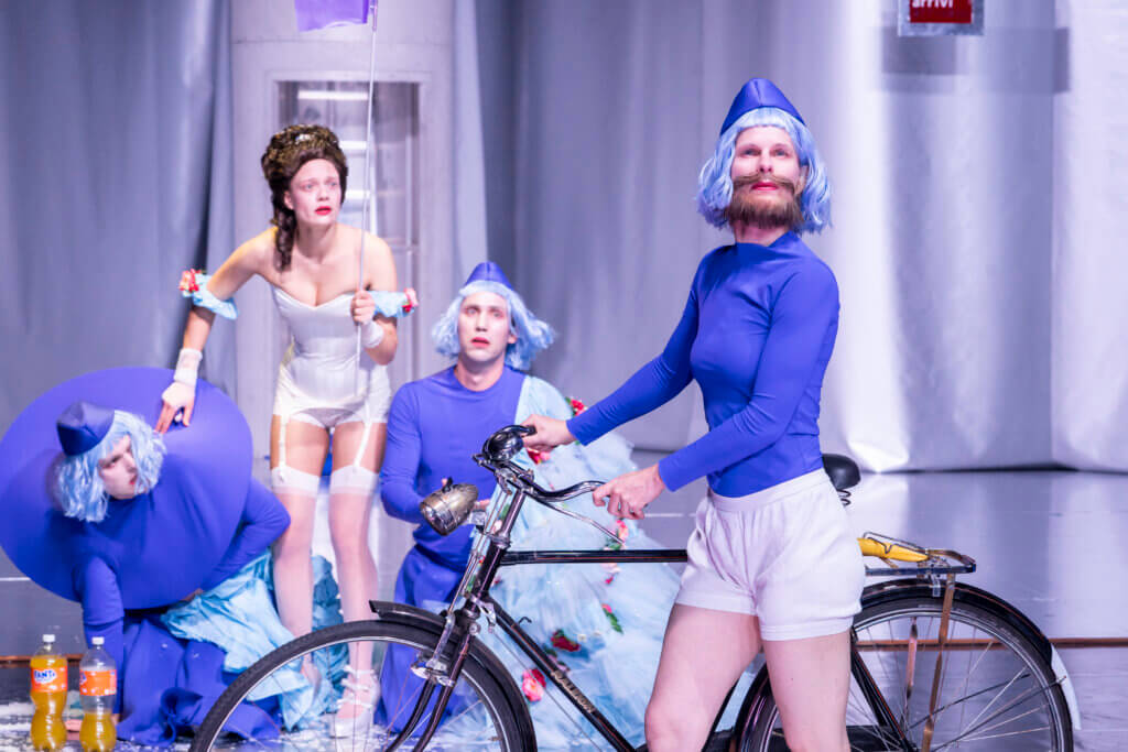 Im Vordergrund steht eine Person in kurzen Bikershorts mit Fahrrad. Dahinter steht eine eine Frau mit eleganter Hochsteckfrisur und Siedenstrümpfen. Ihren Reifrock hält eine witere Person, die neben ihr auf deem Boden kniet.