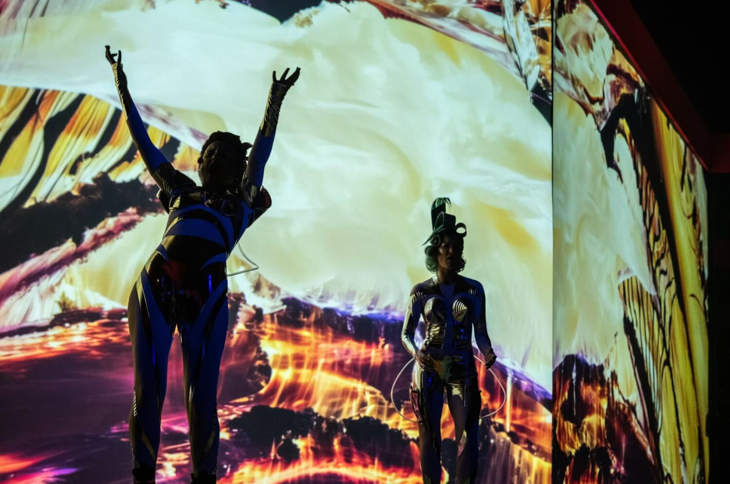 Aufführungsfoto von „KI essen seele auf (Orpheai)“ von Thomas Köck am Schauspiel Stuttgart. Zwei Frauen stehen im Schatten einer riesigen Leinwand, auf der Multimedia projiziert ist.