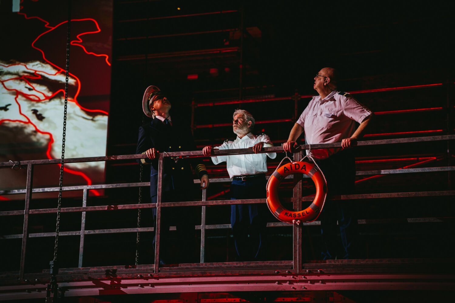 Ian Sidden (Il re dell'Egitto) steht an der Rehling des Kreuzfahrtschiffs Aida und blickt beunruhigt auf das Meer hinaus. Neben ihm stehen zwei Statisten.