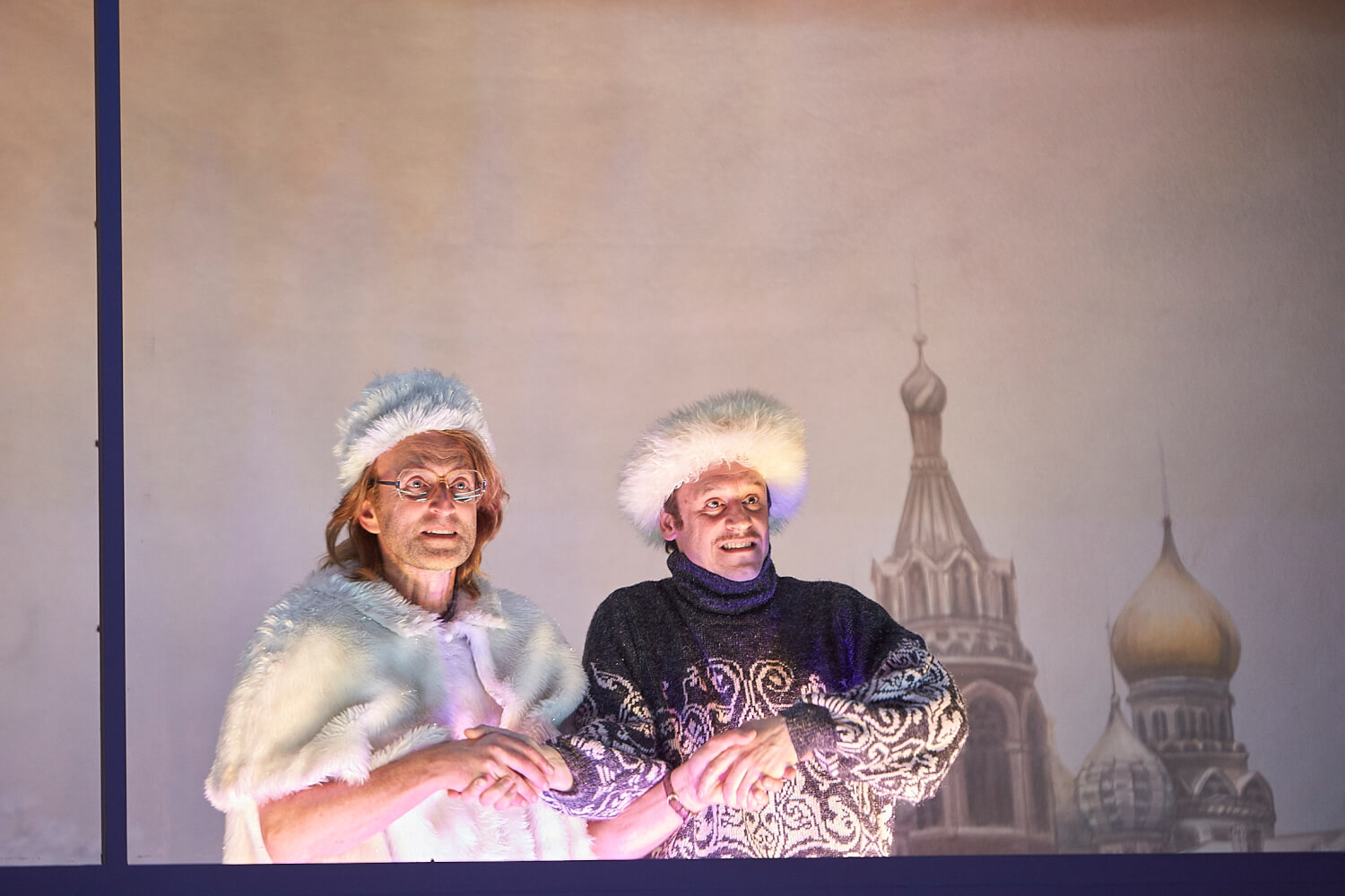 Aufführungsfoto von „Anna Karenina“ mit Clemens Sienknecht und Jan-Peter Kampwirth. Die zwei Männer stehen nebeneinander mit überkreuzten Armen halten sie sich an den Händen und gucken nach vorne. Sie tragen weiße Pelzmützen.