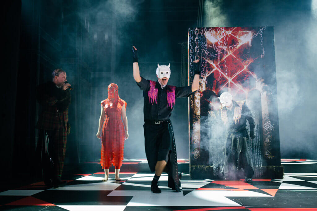 „Meister und Margarita“ von Michail Bulgakow am schauspiel erlangen. Eine Person mit einer weißen Maske mit Hörnern streckt die Zunge raus und hebt die Arme nach oben. Links spricht ein Mann in ein Mikrophon. Zwischen den beiden steht eine Frau in einem roten Kostum, das ihr Gesicht verdeckt. Rechts kommt eine weitere Person mit einer weißen Maske mit Hörnern. Der Boden ist schwarz, rot, weiß gemustert.