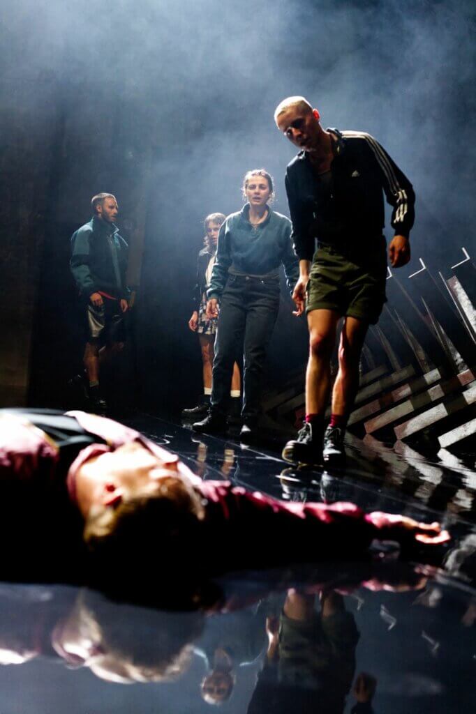 „Das große Heft“ nach dem Buch von Ágota Kristóf am Volkstheater München. Eine Person liegt dicht vor der Kameralinse auf dem Boden, die Arme von aich gestreckt. Vier Personen im Hintergrund schauen sich an. Zwei Personen weiter vorne betrachten den am Boden liegenden Mann.