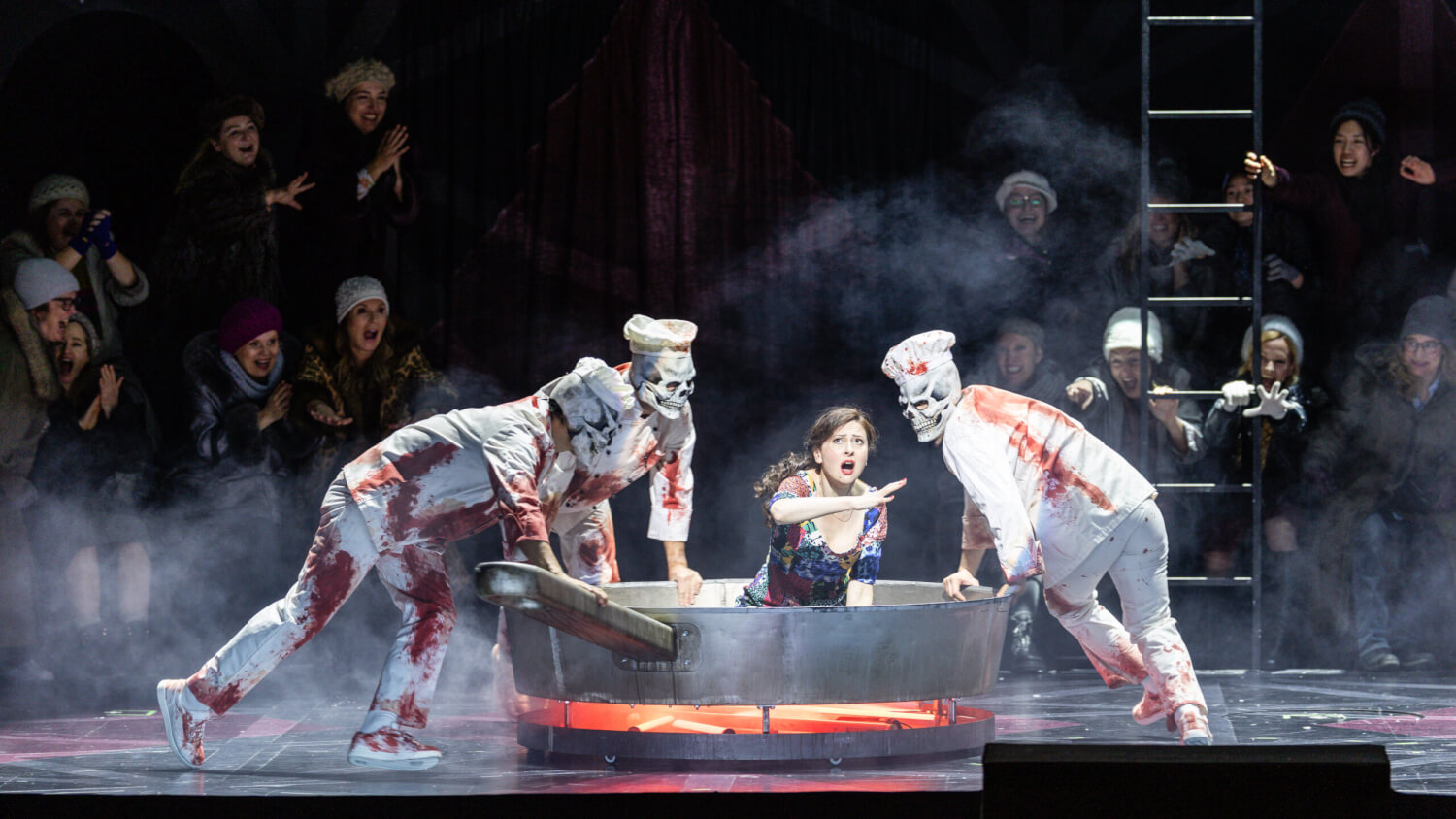 Aufführungsfoto von „Die Nacht vor Weihnachten“ von Nikolai Rimski-Korsakow an der Bayerischen Staatsoper München. Eine Frau sitzt in einer riesigen gusseisernen Bratpfanne. Drei Gestalten mit Totenköpfen und blutverschmierter Kleidung fassen die Pfanne am Rand an und schauen sie an.