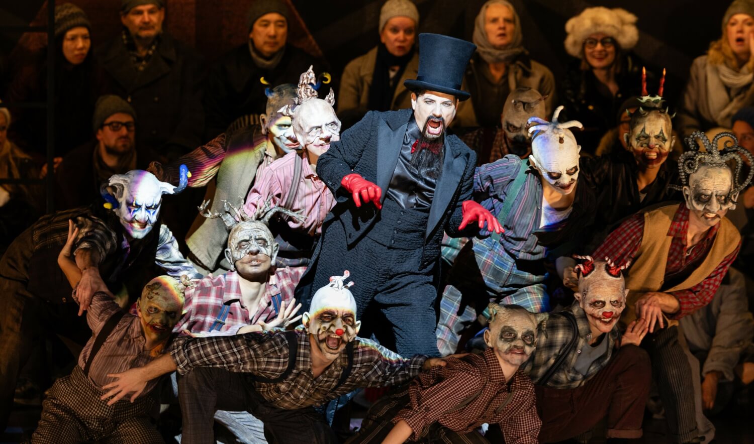 Aufführungsfoto von „Die Nacht vor Weihnachten“ von Nikolai Rimski-Korsakow an der Bayerischen Staatsoper München. Ein Mann in Frack und Zylinder mit roten Handschuhen, steht in einer Gruppe aus Wesen mit unheimlichen Masken.